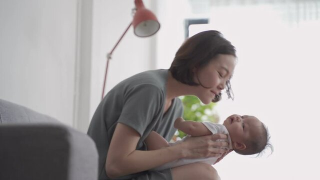 asian woman mother holding happy baby infant playing and kissing on sofa in living room at minimal style home in day time. candid authentic shot life at home. mom play with baby girl by love and care.