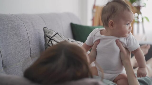 asian woman mother holding happy baby infant playing and kissing on sofa in living room at minimal style home in day time. candid authentic shot life at home. mom play with baby girl by love and care.
