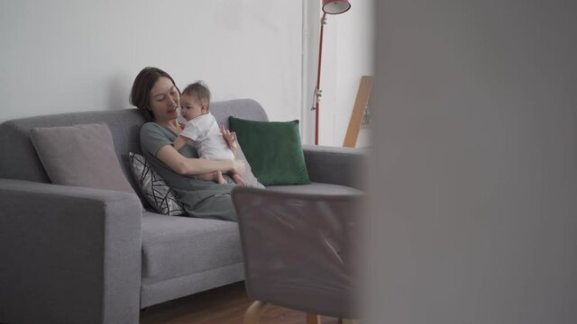 asian woman mother holding happy baby infant playing and kissing on sofa in living room at minimal style home in day time. candid authentic shot life at home. mom play with baby girl by love and care.