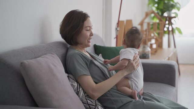 asian woman mother holding happy baby infant playing and kissing on sofa in living room at minimal style home in day time. candid authentic shot life at home. mom play with baby girl by love and care.