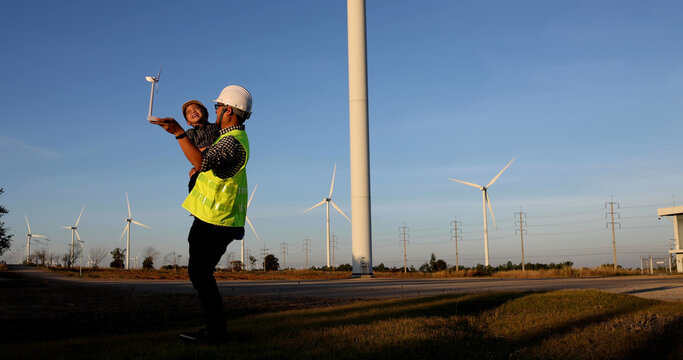 Asian Father Engineer Enjoying And Holding Son In The Wind Turbines Farm, Family With Environmental Engineering Renewable Energy Concept