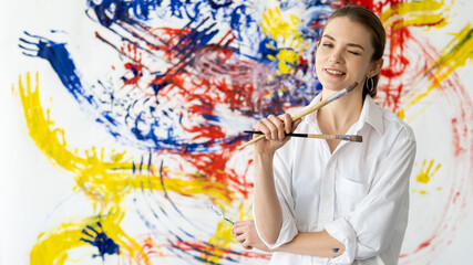 Fun painting. Street art. Creative hobby. Portrait of playful confident artist woman in white with paintbrushes on colorful yellow blue red abstract design wall background.