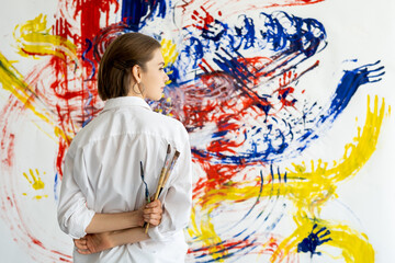 Art therapy. Hand painting. Relaxing hobby. Peaceful pensive painter woman with paintbrushes colorful yellow blue red smeared handprint abstract pattern on white wall.