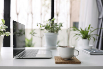 White coffee cup with laptop side view and plant pot on white desk indoor work from home