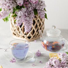 Spring composition with a cup of purple tea, teapot and lilac flowers on light background. Spring tea party, tea drinking. Menu, greeting card. Square