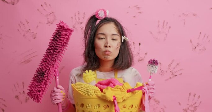 Positive funny Asian woman makes grimace blows cheeks sticks out tongue poses with cleaning equipment foolishes around while tidying up room isolated over pink background. Housework concept.