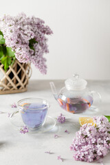 Purple tea in cup, teapot, lemons and a bouquet of blooming lilac on light background. Spring tea drinking. Greeting card, invitation design. Cafe menu, poster. Lilac tea recipe. Side view, copy space