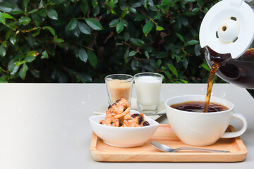 pouring coffee into cup and on the table also have cookies, milk and sugar. Photo taken in the garden scenery.