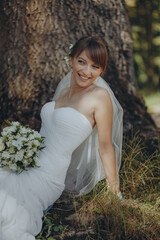 Fototapeta na wymiar Beautiful bride with long veil sitting in a forest, holding bridal bouquet