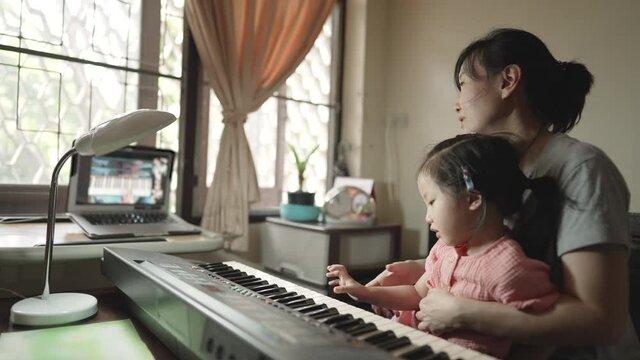 Asian mother helping and teaching daughter to play the piano during music lesson for online learning.