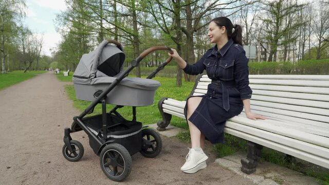 Mother with baby stroller on a walk in spring park, woman in denim dress with baby carriage sits on a bench in the park. High quality 4k footage