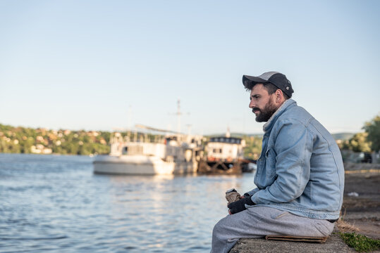 Young depressed suicidal drunk homeless man sitting under the bridge drinking alcohol drink and thinking to jump in to the river to commit suicide and end his life, social documentary concept