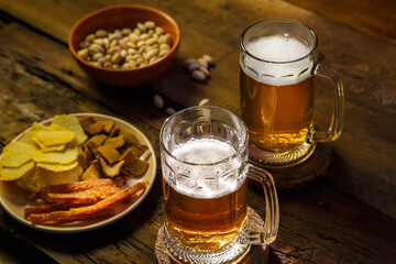 Two mugs with foamy beer and pistachios with chips on a wooden table.
