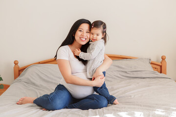 Asian Chinese pregnant woman with toddler girl hugging on bed at home. Girl daughter kid playing...