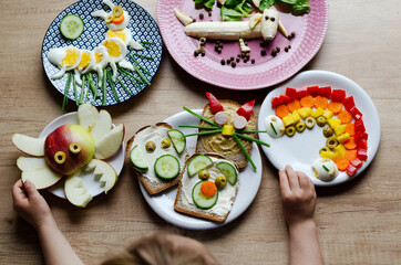 Fun with food with little child. Colorful table with different vegetables and healthy kids food. Hands of child with plates of creative breakfast.