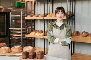Portrait of young woman in apron looking at camera while standing in the bakery with fresh bread in...