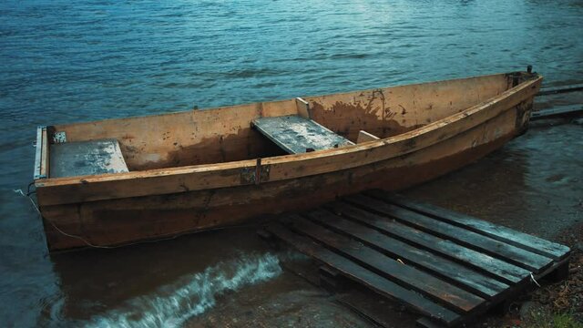 An old makeshift boat made of wood bobs on the waves. It slams its starboard side against the wooden scaffolding. The boat has two seats for passengers. 