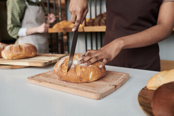 Close-up of worker in apron cutting fresh bread on cutting board at the table