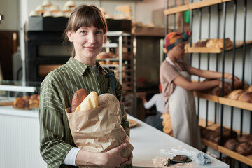 Portrait of young woman looking at camera and holding purchases she buying fresh bread in bakery...