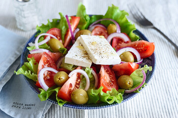 Greek salad of fresh vegetable with tomatoes, lettuce, olives, red onion and feta cheese in bowl on white striped linen tablecloth. Selective focus