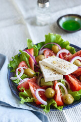 Greek salad of fresh vegetable with tomatoes, lettuce, olives, red onion and feta cheese in bowl on white striped linen tablecloth. Selective focus
