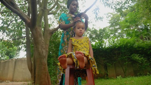 A down low shot of a young African girl in bare feet wearing colorful African clothing as her mother braids her hair in a Afro cultural style.