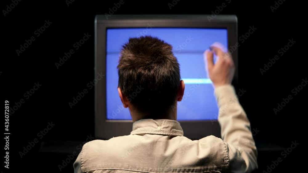 Poster rear view of a man sitting in front of an old tv with noise interference on a blue tv monitor in a d