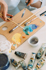 Painting supplies. Art hobby. Creative occupation. Professional female artist hands mixing yellow blue color oil or acrylic paint from tubes with paintbrush knife on wooden palette.