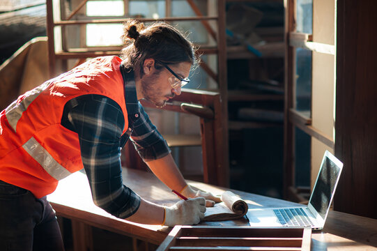 Young and handsome caucasian carpenter in the carpentry shop is working and checking blueprint in laptop to repair a furniture. - Powered by Adobe