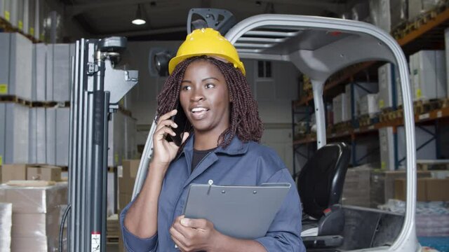 Excited female factory employee in helmet talking on phone. Smiling African American worker standing in storehouse, discussing order with supplier. Warehouse, telephone communication concept.