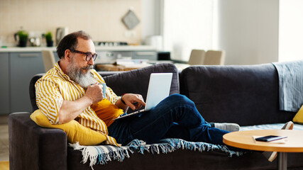 Side view of senior man with gray beard and in horn-rimmed eyeglasses working on laptop computer and drinking tea sitting on comfortable sofa at home