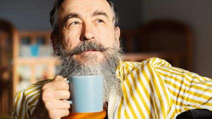 Senior man with gray beard and mustache drinking tea or coffee, looking away and thinking or day dreaming  sitting at home in morning..