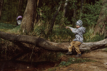 cute little caucasian boy sitting on the log over the stream with long twig pretending its a fishing rod. Image with selective focus.