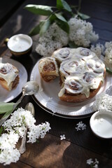 Cinnamon rolls covered with butter cream on a table with white lilacs. Breakfast with rolls and milk.