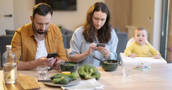Young parents sitting in smart phones during a lunch time at the table with their one year baby. Dependence of parents on mobile devices and not paying attention to their children