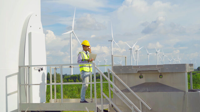Portrait of Asian windmill engineer man, worker working, using a computer laptop on site at wind turbines field or farm, clean energy source. Eco technology for electric. industry environment. People