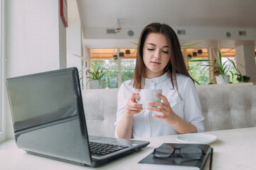 A girl in a white shirt sits at a table in a cafe, holds a cup of coffee.