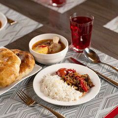 Set lunch from rice with vegetables, soup with meat broth, samsa and red compote on blurred background. Beautifully served dishes with delicious hearty food on wooden table with cutlery on it.