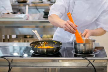 Chef hands holding silicone spatula and cooking vegetables in pan on electric stove - front view. Professional cooking, cookery, gastronomy and food concept