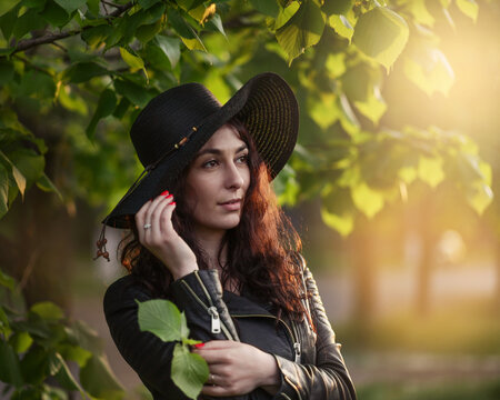 Close up portrait of Beautiful girl in dark clothet and black hat standing near green leaves on sunset. Art work of romantic woman.