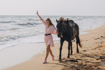A young beautiful girl in a pink dress walks and plays with a black horse on the seashore. Holiday,...