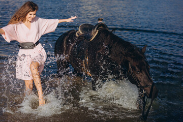 A young beautiful girl in a pink dress walks and plays with a black horse on the seashore. Holiday, fun, vacation