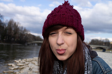 Porträt junge freche Frau am Fluss mit roter Mütze im Herbst steckt ihre Zunge raus