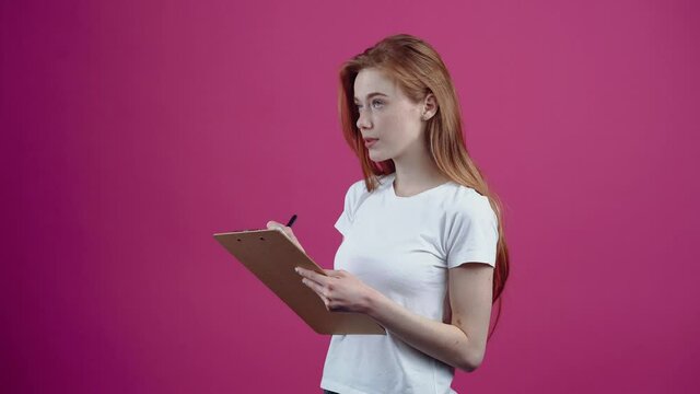 Portrait of an entrepreneur who makes the to-do list and thinks about what else to add. Freckled teenager in a white T-shirt, isolated on a pink background. The concept of people's lifestyle
