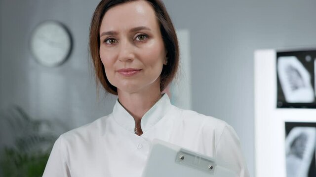Portrait Of White Female Doctor In White Coat. Woman Standing In The Office Holding Notebook.