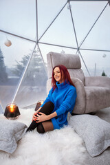 beautiful young girl with red hair and a blue sweater sits near a gray armchair in the living room by the panoramic window