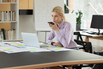 Confident businesswoman recording audio voice message on smartphone sitting at table with laptop and documents in office room, chatting with colleagues online or speakerphone in social networks
