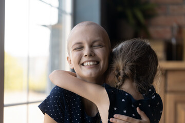 Overjoyed young mother with cancer hugging daughter girl. Happy ill hairless mom enjoying leisure...