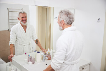 Obraz premium Happy smiling adult male looking to the reflection in the mirror in bathroom
