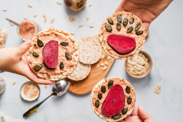 Fototapeta na wymiar Crispy puffed rice cakes on table hummus spread and tomato and beet vegetables and sesame pumpkin seeds on the table - top view on hands holding healthy vegetarian or vegan breakfast gluten free